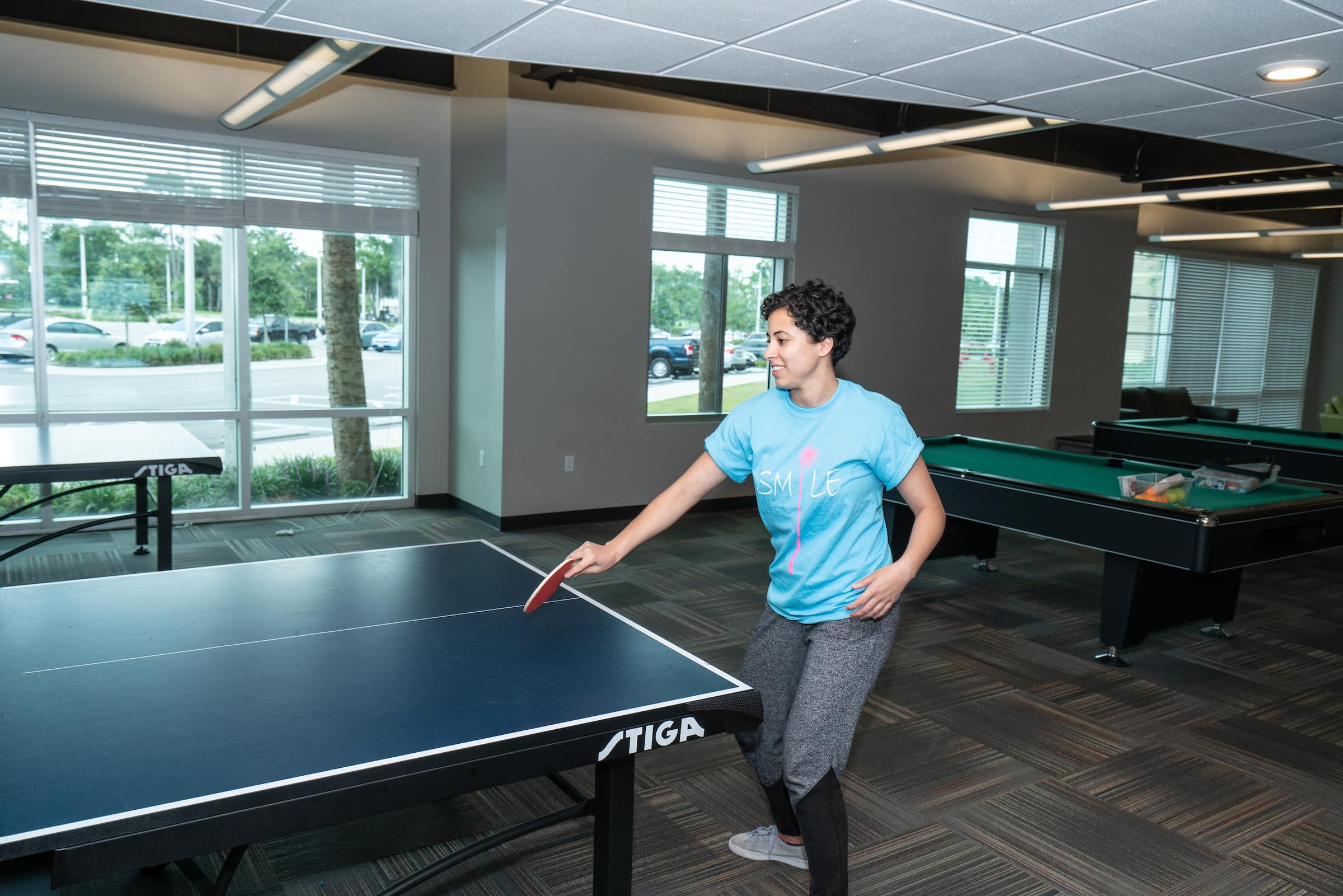 A student plays ping pong with a pool table in the background in New hall.