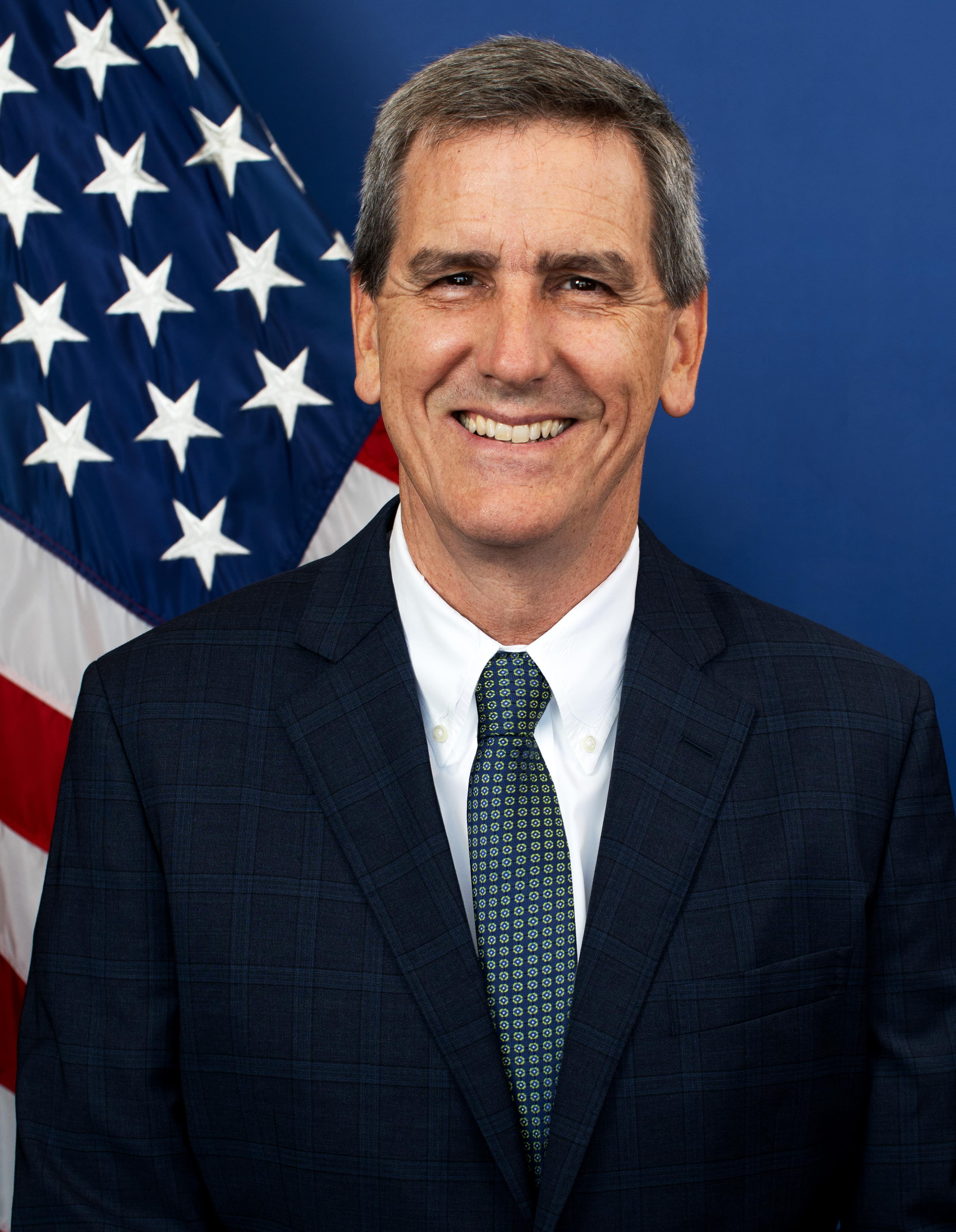A headshot of Michael Whitaker wearing a suit and tie.  There is an American flag behind Michael.