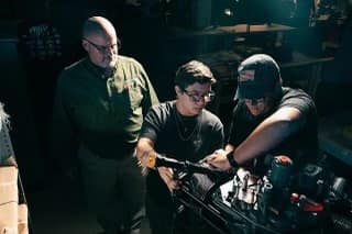 A professor supervising two students working on an engine.