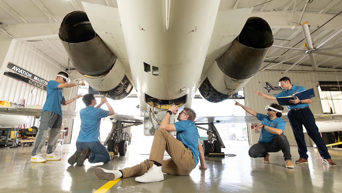 Five students in blue shirts inspect the underside of a jet aircraft